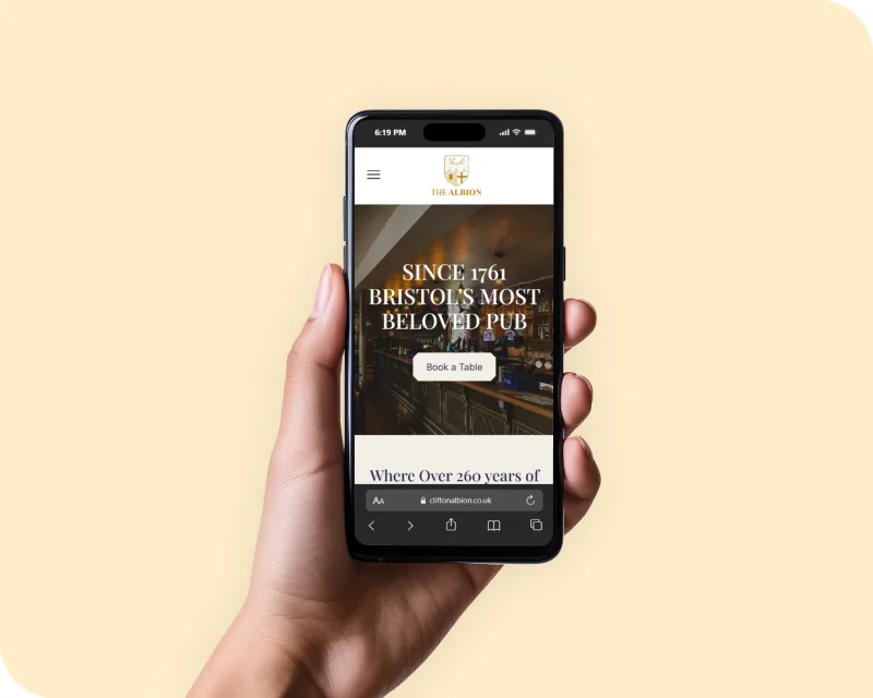 Hand holding a smartphone showing The Albion Bristol homepage hero with the headline "Since 1761 Bristol's Most Beloved Pub" and a book a table call to action button on a warm yellow background