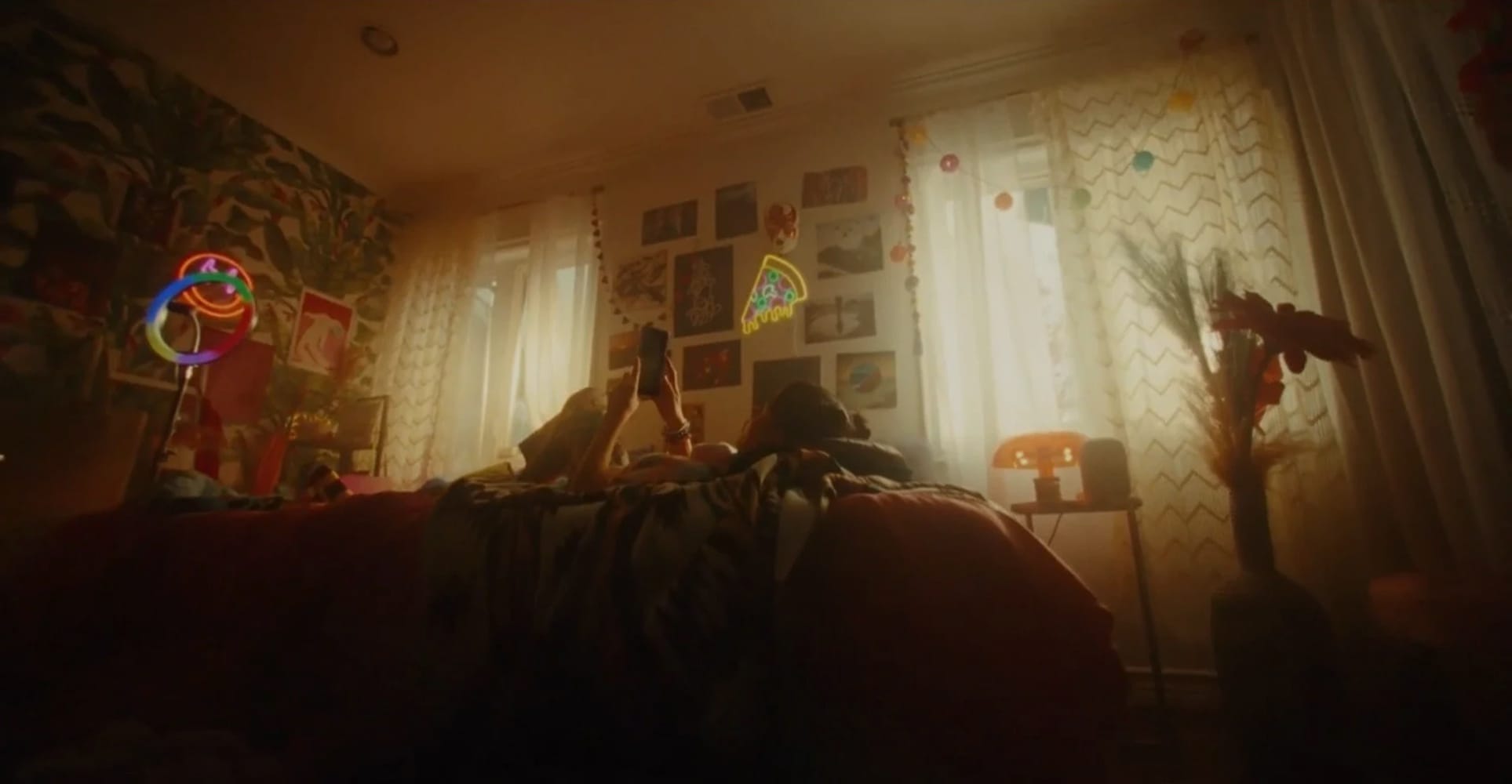 A person lying on a bed in a warmly lit, eclectic bedroom holding a phone, with neon light props and a gallery wall in the background, captured during an Imagine This production