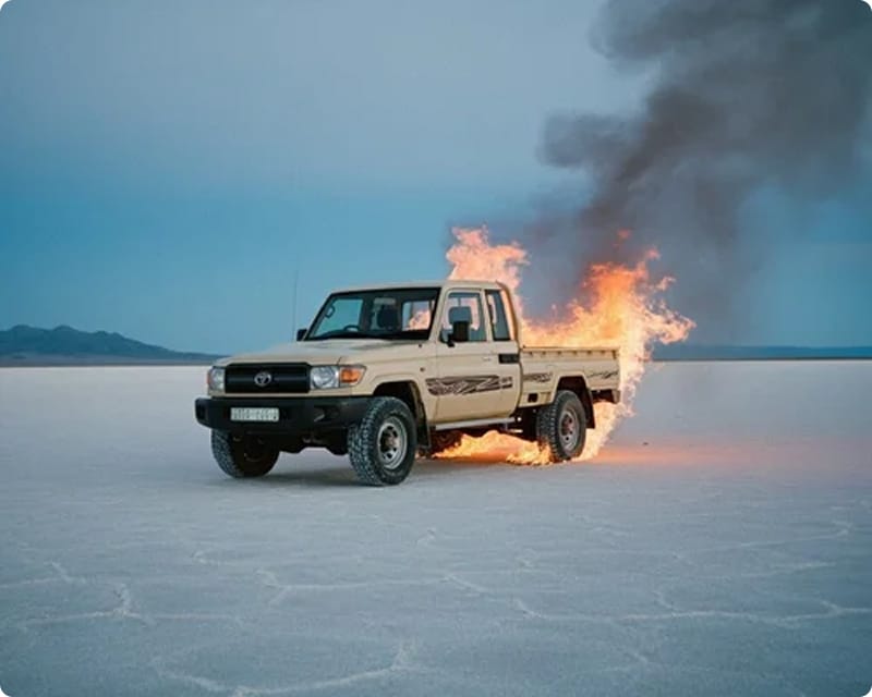 A pick-up truck engulfed in flames on a vast salt flat with dark smoke rising against an overcast sky, captured during a film production shoot